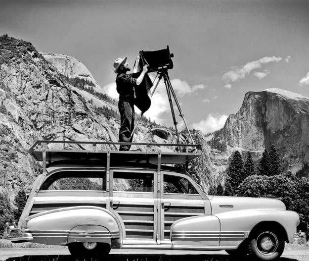 Ansel Adams standing on his 1947 Pontiac Streamliner using a large format camera